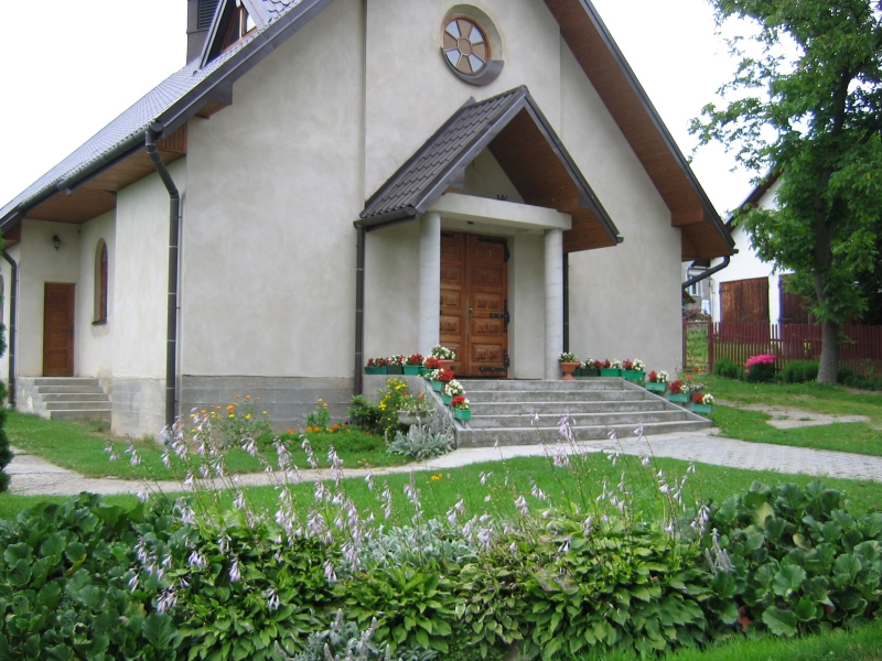 Kaplica w Boguszowej, niewielki budynek ze spadzistym dachem, przy wejściu dwie kulumny, wejście przykryte daszkiem, nad wejściem okrągłe okno z witrażem w kształcie krzyża, przed budynkiem chodnik i trwanik z krzewami i kwiatami