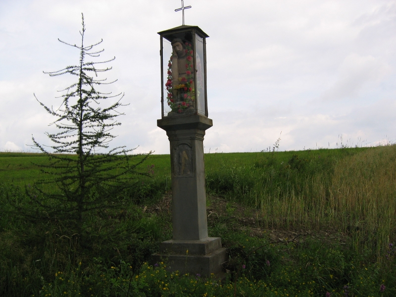 Przydrożna kapliczka - figura Chrystusa na postumencie, na górze krzuż, figura ozdobiona kwiatami, obok postumentu choinka a w tle łąka