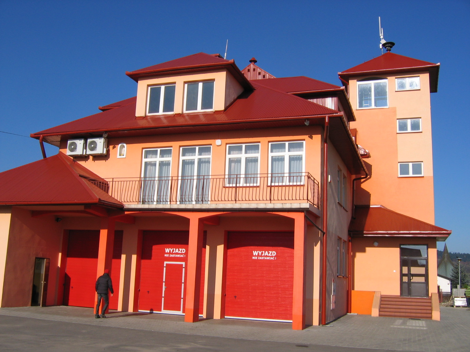 Budynek remiozy strażackiej w Niskowej - pomarańczowy 2 piętrowy budynek ze spadzistym czerwonym dachem. Na zdjęciu widoczne 3 czerwone bramy garażowe oraz wejście do budynku, po prawej stronie budynku widoczna wieża obnserwacyjna, przed budynkiem podjazd, w tle niebieskie niebo