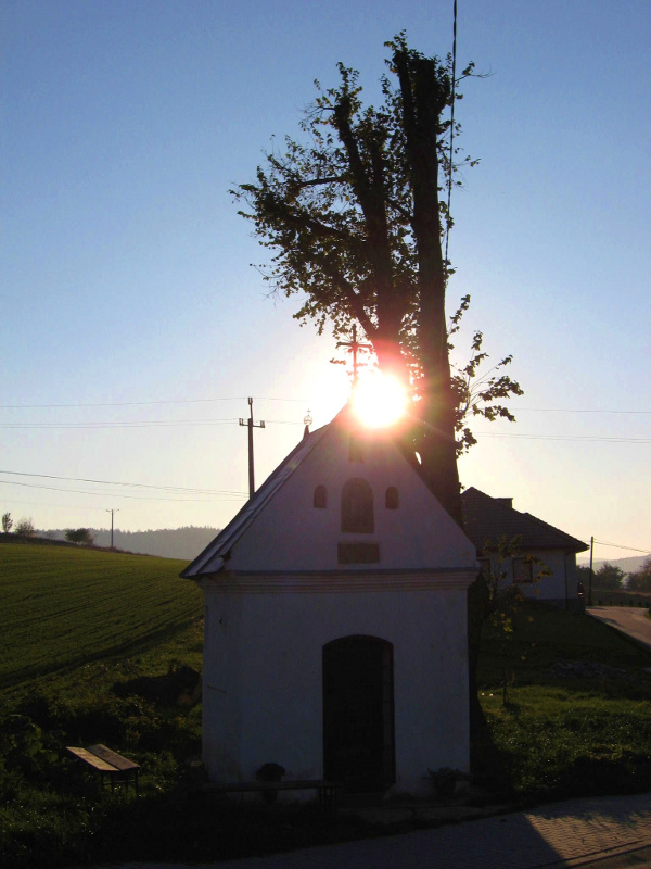 Przydrożna kapliczka z trójkątnym dachem, w tle łąka oraz słońce chowające się za dachem kapliczki