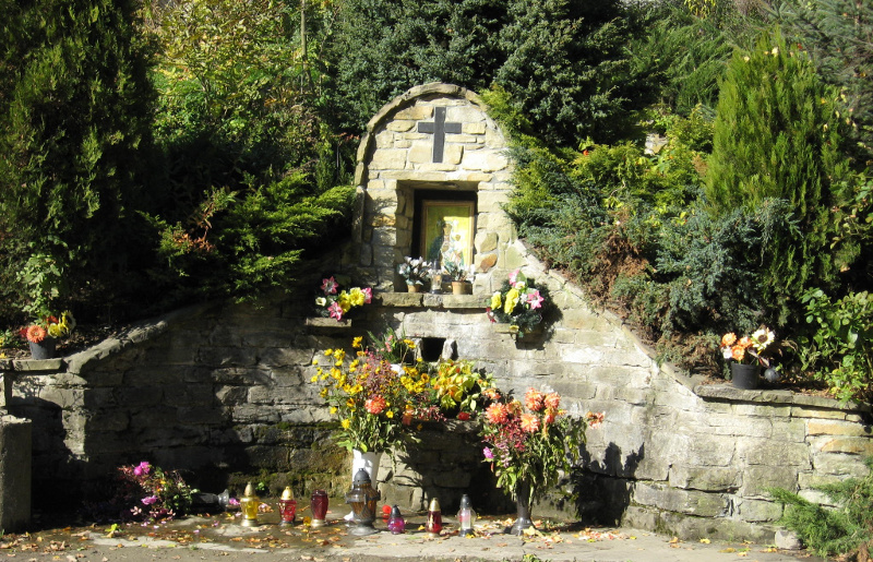 Kapliczka murowana z krzyżem wkomponowana w zbocze, otoczona roślinnością i kwiatami w doniczkach i flakonach