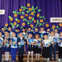 Dzieci odświętne ubrane ustawione w dwóch rzędach do wspólnego zdjęcia - w tle niebieska kotara udekorowana słonecznikami oraz napis ŚLUBOWANIE