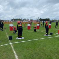 Zawody strażackie na boisku sportowym - grupy strażaków celują wężami strażackimi w wyznaczony punkt