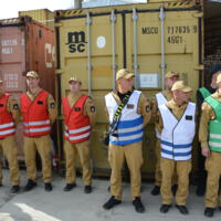 Osoby ustawione w rzędzie w szarych i niebieskich mundurach niektóre w odblaskowych kamizelkach, w tle czerwone i szare kontenery