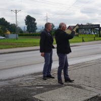Wójt gminy Bernard Stawiarski i Starosta Powiatu Nowosądeckiego stoją obok siebie na drodze,  starosta ma uniesioną rękę i na coś wskazuje, w tle droga i budynki mieszkalne