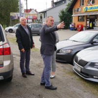 Wójt gminy Bernard Stawiarski i Starosta Powiatu Nowosądeckiego oraz dyrektor ZGKiM w Chełmcu  stoją obok siebie na parkingu,  wójt ma uniesioną rękę, w tle droga i budynki mieszkalne