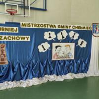 Na sali gimnastycznej tablica z napisem Mistrzostwa Gminy Chełmiec Turniej Szachowy - w rogu Herb gminy, niebieskie tło rysunek z szachami
