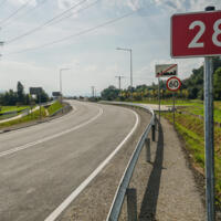 Zdjęcie drogi z widocznym znakiem z numerem 28 na czerwonym tle - w dalszej perspektywie znak ograniczenia prędkości do 60 oraz koniec obszaru zabudowanego, nad drogą linia energetyczna