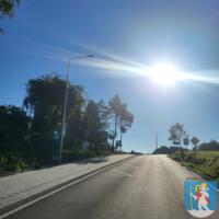 Asfaltowa droga - po lewej stronie chodnik, pobocza porośnięte trawą i drzewami, w oddali domu, na niebieski niebie słońce