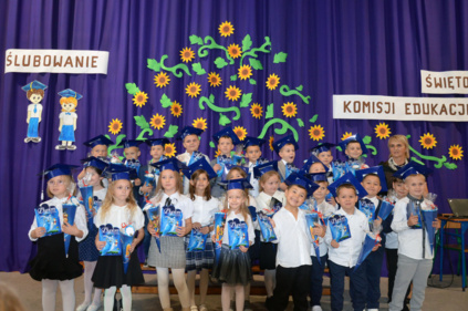 Dzieci odświętne ubrane ustawione w dwóch rzędach do wspólnego zdjęcia - w tle niebieska kotara udekorowana słonecznikami oraz napis ŚLUBOWANIE
