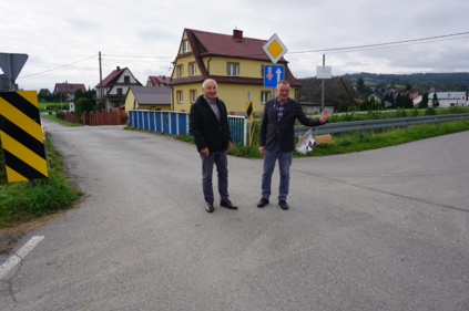 Wójt gminy Bernard Stawiarski i Starosta Powiatu Nowosądeckiego stoją obok siebie na drodze, wójt ma uniesioną rękę, w tle droga i budynki mieszkalne