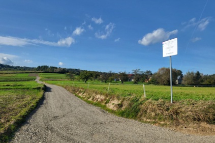 Droga gruntowa - po obu stronach zielone łąki po prawej stronie tabliczka informacyjna