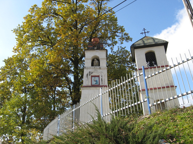 Za ogrodzeniem dwa budynki, po prawej stronie dzwonnica, po lewej kapliczka, w tle drzewa i niebieskie niebo